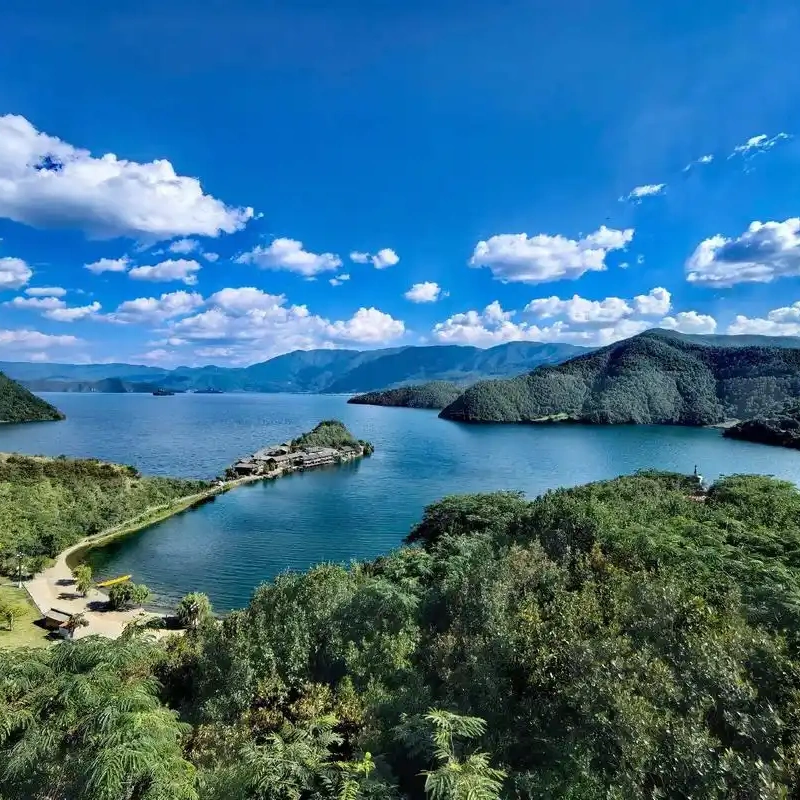 Panoramic view of Lugu Lake 瀘沽湖壯麗湖景在晴朗藍天下展開,湖水深藍清澈,群山環繞湖泊。湖中央的小島與連接岸邊的堤道清晰可見,周圍綠意盎然的森林與遠方山巒層層延伸,白雲點綴天空,展現瀘沽湖高原湖泊寧靜而壯觀的自然風光。