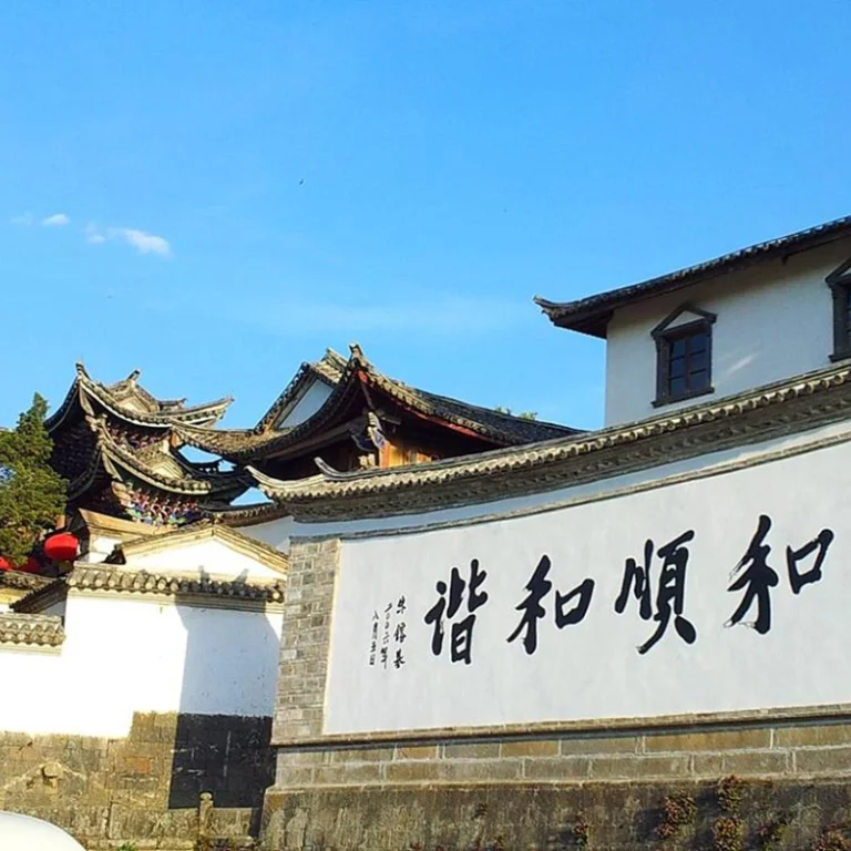 tengchong 雲南騰衝的 和順古鎮 古建築景觀。白色石牆上寫著「和順和諧」書法字樣,牆後可見傳統中式屋頂與飛簷建築,青瓦層疊,展現典型的古鎮風格。晴朗藍天下,古鎮街巷寧靜雅致,整體畫面呈現濃厚的歷史文化氣息與雲南傳統人文風貌。